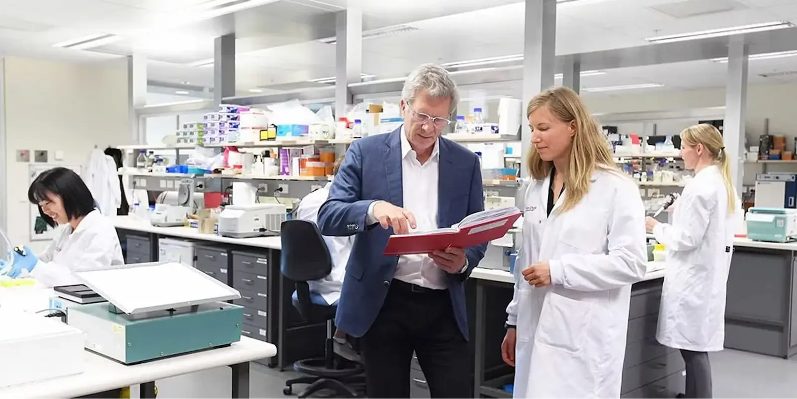 Researchers working in a modern laboratory; a man in a suit reviews a document with a scientist in a lab coat while other lab technicians work in the background.